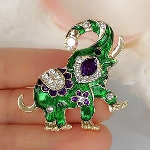 May include: An elephant-shaped brooch featuring vibrant green enamel, gold-tone metal edges, and purple floral accents. The design is embellished with clear, sparkling rhinestones along the trunk, back, and legs, with a large purple stone.