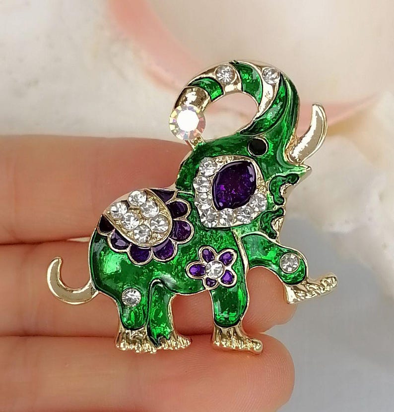 May include: An elephant-shaped brooch featuring vibrant green enamel, gold-tone metal edges, and purple floral accents. The design is embellished with clear, sparkling rhinestones along the trunk, back, and legs, with a large purple stone.