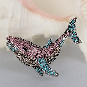 May include: A bejeweled whale brooch in silver-tone metal. The whale is encrusted with pink, brown, and turquoise rhinestones. The brooch features a detailed design of a whale with a tail and fins. The whale's eye is a small black stone.