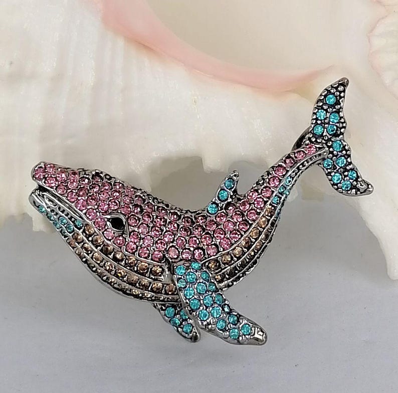 May include: A bejeweled whale brooch in silver-tone metal. The whale is encrusted with pink, brown, and turquoise rhinestones. The brooch features a detailed design of a whale with a tail and fins. The whale's eye is a small black stone.