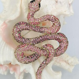 May include: A pink and gold snake brooch, adorned with pink crystals. The snake is coiled, featuring a black head and a pin fastening. The brooch is displayed against a white backdrop.