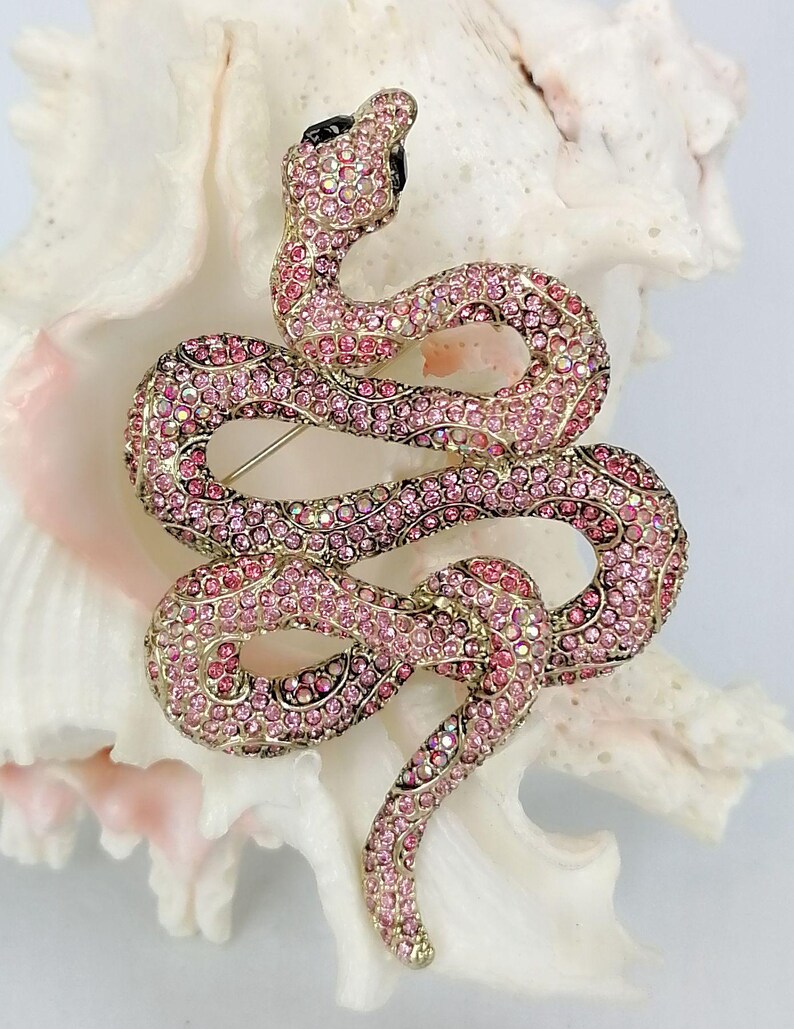 May include: A pink and gold snake brooch, adorned with pink crystals. The snake is coiled, featuring a black head and a pin fastening. The brooch is displayed against a white backdrop.