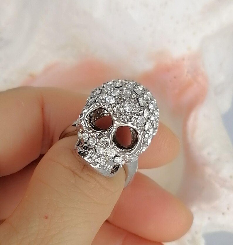 Op de afbeelding: Een zilverkleurige ring met een driedimensionaal schedelontwerp. Het gehele oppervlak van de schedel is ingelegd met talloze kleine, heldere, sprankelende strass-steentjes, terwijl de oogkassen hol en donker blijven, wat zorgt voor een opvallend contrast.