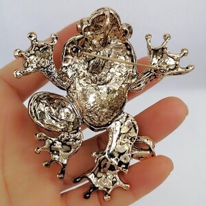 K&ouml;nnte beinhalten: Goldfarbene Froschbrosche mit strukturierter Oberfl&auml;che. Die Brosche zeigt ein detailliertes Design einer Froschfigur mit ausgestreckten Gliedma&szlig;en und einer Nadel auf der R&uuml;ckseite zum Befestigen. Die Augen und die Haut des Frosches sind aufwendig detailliert.