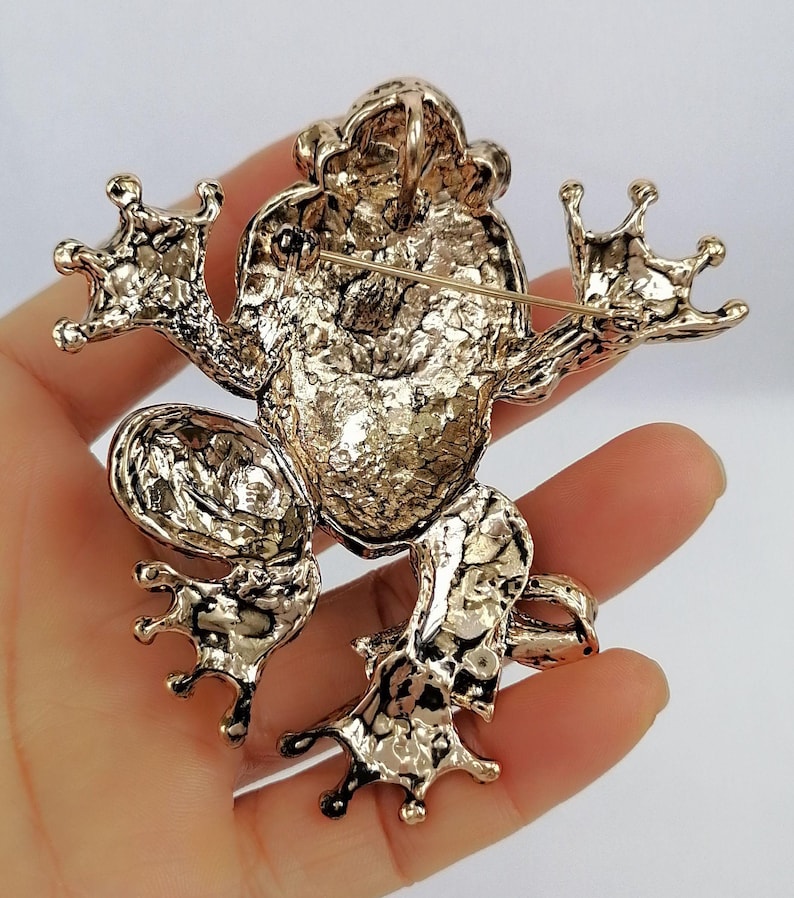 K&ouml;nnte beinhalten: Goldfarbene Froschbrosche mit strukturierter Oberfl&auml;che. Die Brosche zeigt ein detailliertes Design einer Froschfigur mit ausgestreckten Gliedma&szlig;en und einer Nadel auf der R&uuml;ckseite zum Befestigen. Die Augen und die Haut des Frosches sind aufwendig detailliert.