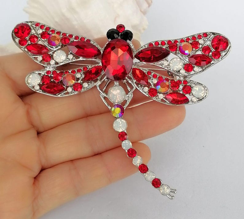 May include: A dragonfly brooch with a silver-tone metal frame. The wings are decorated with red and white gemstones, and the body features a large red gemstone. The dragonfly's head is accented with black gemstones.
