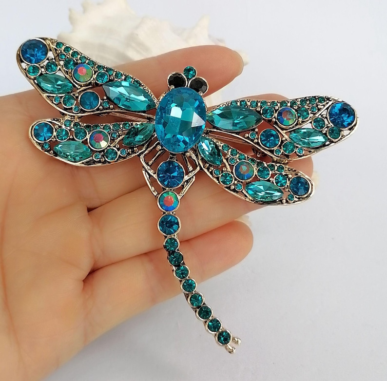 May include: A dragonfly brooch with a gold-coloured metal frame, embellished with turquoise and teal gemstones. The brooch features a large central oval gem, with smaller gems forming the body and wings. The brooch is approximately 10 cm in length.