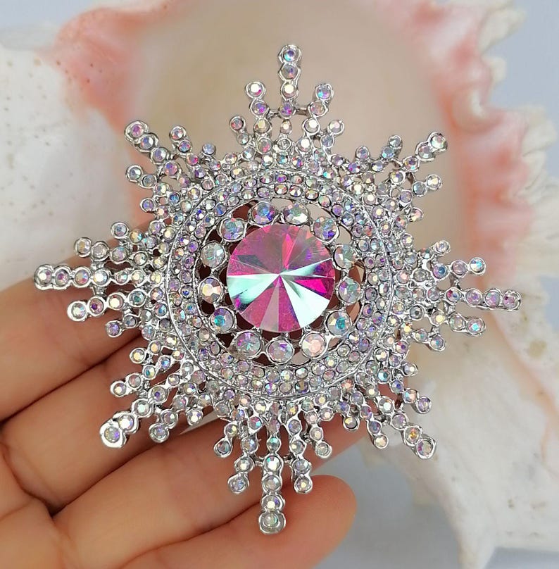 K&ouml;nnte beinhalten: Eine silberfarbene, sternf&ouml;rmige Brosche mit einem gro&szlig;en, runden, irisierenden rosa-blauen Mittelstein, umgeben von zahlreichen kleinen, klaren, funkelnden Kristallen, die in einem strahlenf&ouml;rmigen, schneeflockenartigen Muster angeordnet sind.