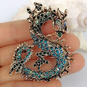 Puede incluir: Broche de drag&oacute;n de estilo antiguo. El drag&oacute;n est&aacute; adornado con pedrer&iacute;a turquesa y verde, engastada en metal dorado. El dise&ntilde;o del drag&oacute;n presenta escamas detalladas y un cuerpo enroscado, creando un accesorio llamativo.