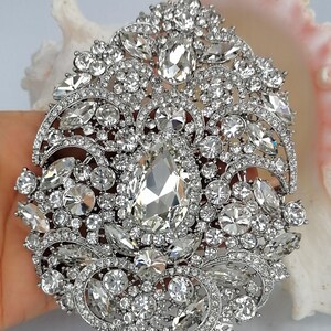May include: A large, ornate silver brooch with a dazzling array of clear, faceted crystals. The brooch features a central, teardrop-shaped crystal surrounded by smaller, round and oval stones. The design is intricate and detailed, with a vintage or antique aesthetic.