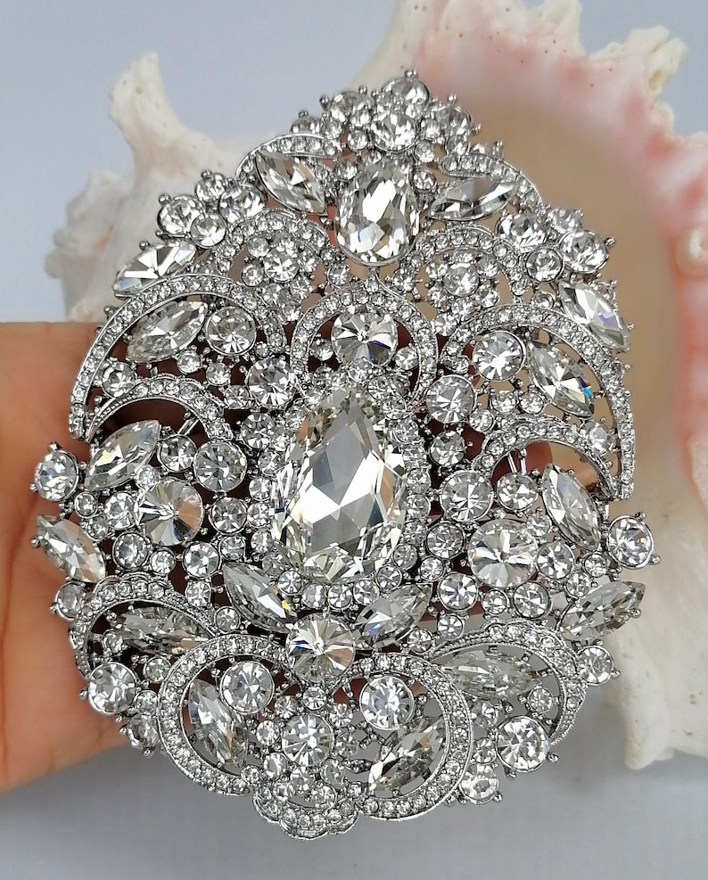 May include: A large, ornate silver brooch with a dazzling array of clear, faceted crystals. The brooch features a central, teardrop-shaped crystal surrounded by smaller, round and oval stones. The design is intricate and detailed, with a vintage or antique aesthetic.