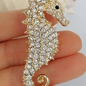 May include: A gold-toned seahorse brooch encrusted with clear, sparkling rhinestones. The seahorse has a detailed design with a curled tail and a small black eye. This elegant accessory is perfect for adding a touch of marine-inspired glamour to any outfit.