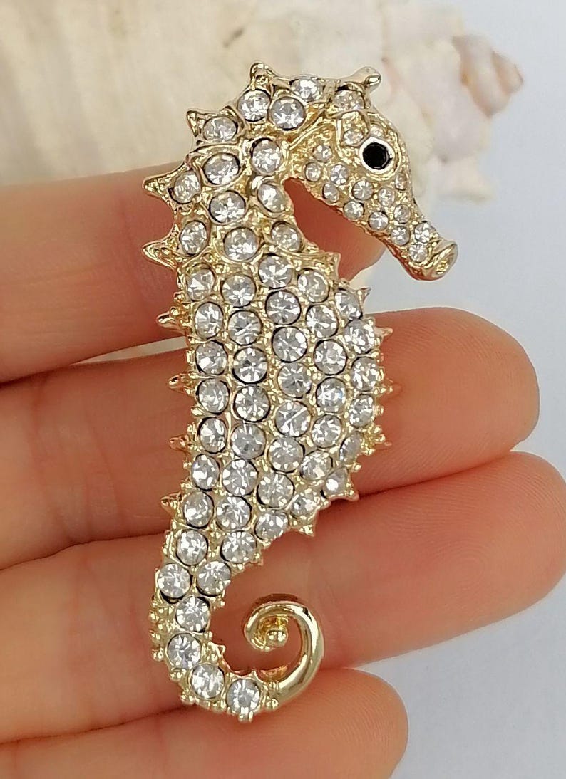 May include: A gold-toned seahorse brooch encrusted with clear, sparkling rhinestones. The seahorse has a detailed design with a curled tail and a small black eye. This elegant accessory is perfect for adding a touch of marine-inspired glamour to any outfit.