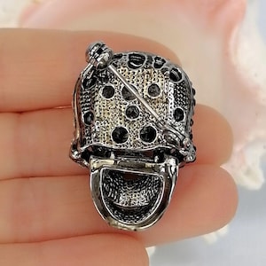 Pode incluir: Um broche de metal prateado com uma parte superior abobadada e texturizada com recortes circulares e uma se&ccedil;&atilde;o inferior lisa e curva. A parte de tr&aacute;s do broche inclui um mecanismo de alfinete horizontal com um fecho girat&oacute;rio.