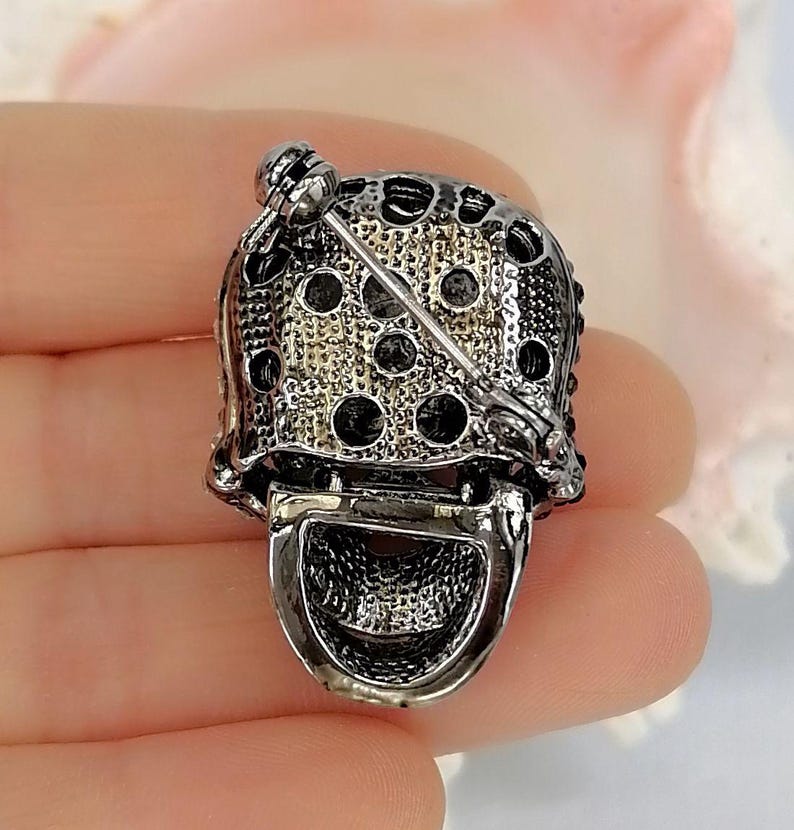 Pode incluir: Um broche de metal prateado com uma parte superior abobadada e texturizada com recortes circulares e uma se&ccedil;&atilde;o inferior lisa e curva. A parte de tr&aacute;s do broche inclui um mecanismo de alfinete horizontal com um fecho girat&oacute;rio.