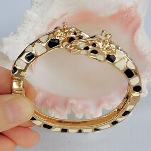 May include: A gold-tone hinged bangle bracelet featuring two giraffe heads meeting at the center. The bracelet is decorated with an enamel giraffe print pattern in black and cream colors, covering the entire surface of the metal band.