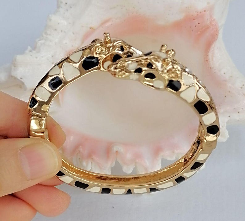 May include: A gold-tone hinged bangle bracelet featuring two giraffe heads meeting at the center. The bracelet is decorated with an enamel giraffe print pattern in black and cream colors, covering the entire surface of the metal band.