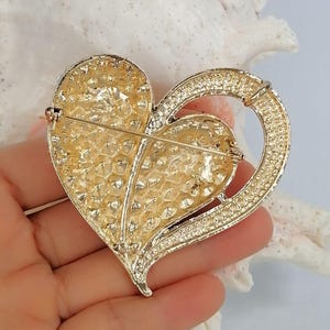 May include: A gold-coloured heart-shaped brooch with a textured surface and a ring detail. The brooch has a pin closure on the back. The heart is embellished with small, sparkling accents.