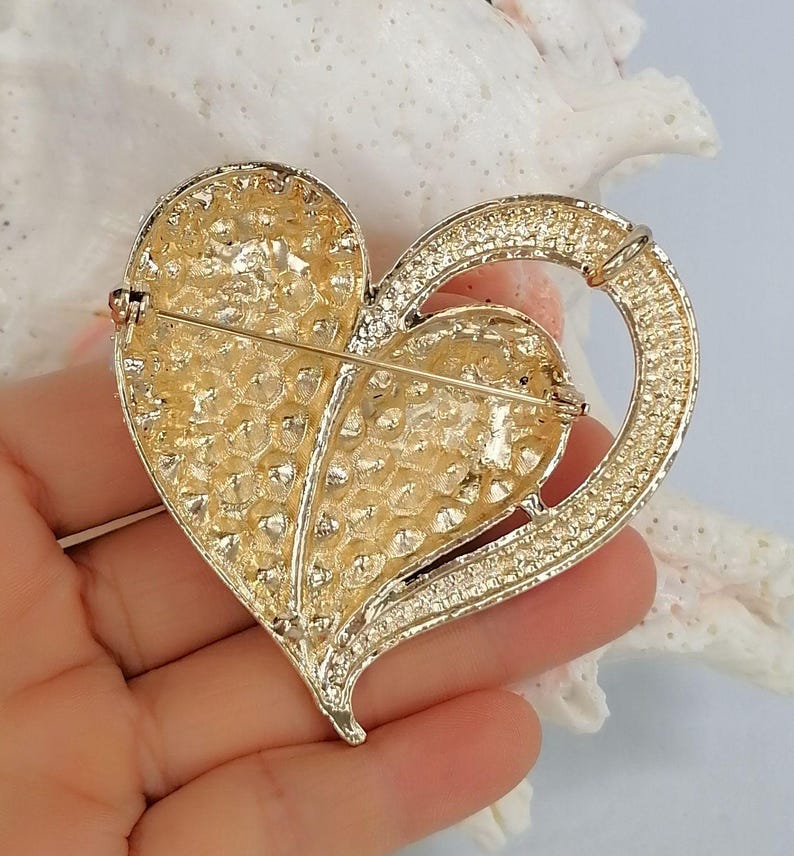 May include: A gold-coloured heart-shaped brooch with a textured surface and a ring detail. The brooch has a pin closure on the back. The heart is embellished with small, sparkling accents.