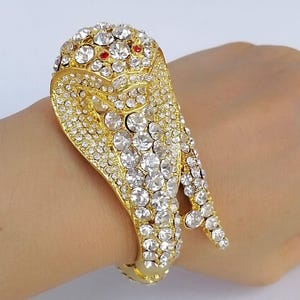 Può includere: Un bracciale rigido in metallo dorato a forma di serpente cobra, caratterizzato da una superficie testurizzata interamente incastonata con strass trasparenti, rotondi e sfaccettati, e due piccoli strass rossi circolari come occhi.