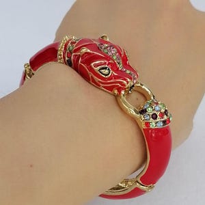 Può includere: Un bracciale rigido incernierato color oro con un design a testa di pantera stilizzata. Il bracciale è rifinito con smalto rosso lucido e impreziosito da strass multicolori incastonati lungo la testa della pantera e su una chiusura circolare.