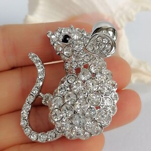 May include: A silver-toned mouse brooch, encrusted with clear, sparkling crystals. The brooch features a detailed design with a rounded body, a long curved tail, and a black eye. The mouse's ear is a teardrop shape.