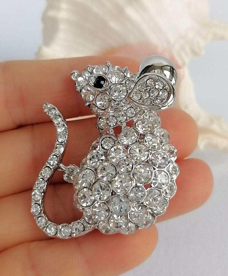 May include: A silver-toned mouse brooch, encrusted with clear, sparkling crystals. The brooch features a detailed design with a rounded body, a long curved tail, and a black eye. The mouse's ear is a teardrop shape.