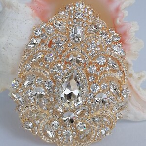 May include: A gold-toned brooch with a complex, symmetrical design. The brooch is encrusted with numerous clear, sparkling crystals of various shapes and sizes, including a large, teardrop-shaped central stone. The brooch is set against a seashell background.