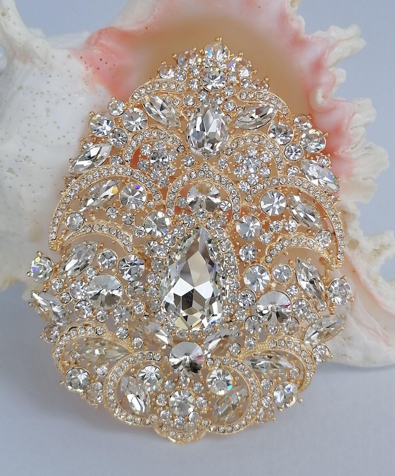 May include: A gold-toned brooch with a complex, symmetrical design. The brooch is encrusted with numerous clear, sparkling crystals of various shapes and sizes, including a large, teardrop-shaped central stone. The brooch is set against a seashell background.