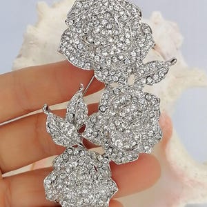 May include: A silver-toned brooch featuring three rose blossoms and two leaves, entirely encrusted with clear, sparkling rhinestones. The floral arrangement is connected by a metal stem, creating a cohesive, shimmering botanical design.