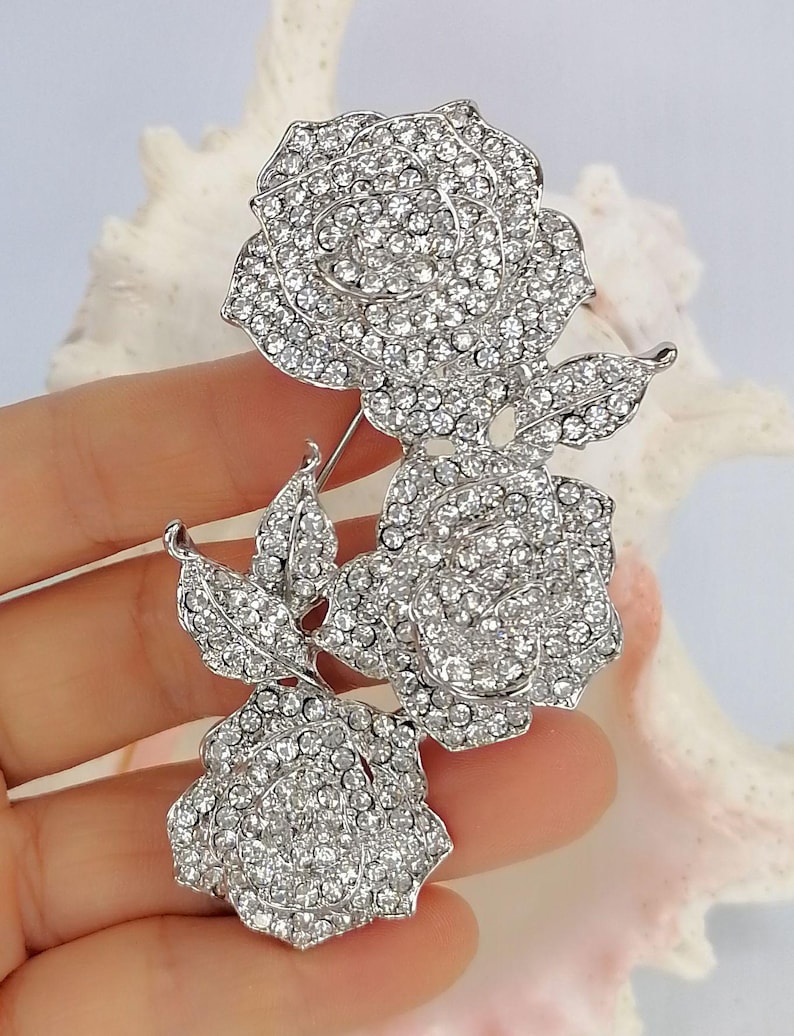 May include: A silver-toned brooch featuring three rose blossoms and two leaves, entirely encrusted with clear, sparkling rhinestones. The floral arrangement is connected by a metal stem, creating a cohesive, shimmering botanical design.