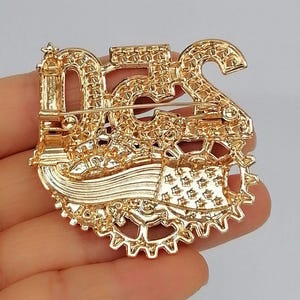 May include: Gold-toned brooch with the number "52" and a gear design. The brooch has a pin on the back for secure attachment. The brooch is held in a hand.