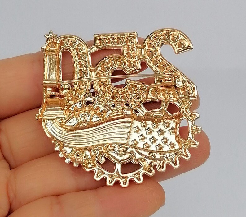May include: Gold-toned brooch with the number "52" and a gear design. The brooch has a pin on the back for secure attachment. The brooch is held in a hand.