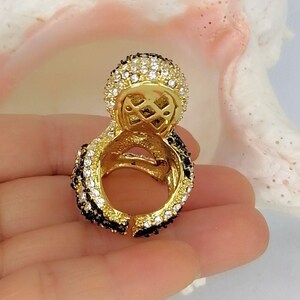 Peut inclure: Une bague dor&eacute;e dot&eacute;e d'une partie sup&eacute;rieure circulaire pro&eacute;minente incrust&eacute;e de cristaux blancs transparents. L'anneau et le sertissage circulaire sont orn&eacute;s de rang&eacute;es altern&eacute;es de cristaux noirs et blancs, cr&eacute;ant un contraste marqu&eacute;.