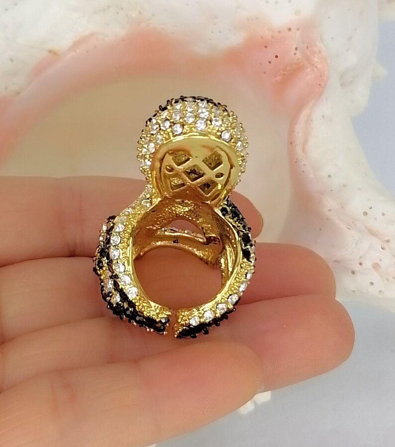 Peut inclure: Une bague dor&eacute;e dot&eacute;e d'une partie sup&eacute;rieure circulaire pro&eacute;minente incrust&eacute;e de cristaux blancs transparents. L'anneau et le sertissage circulaire sont orn&eacute;s de rang&eacute;es altern&eacute;es de cristaux noirs et blancs, cr&eacute;ant un contraste marqu&eacute;.