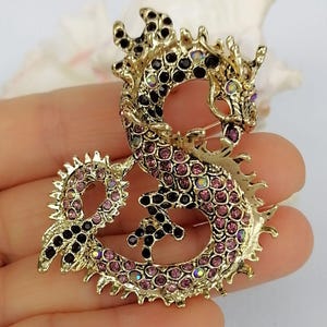 Puede incluir: Broche de drag&oacute;n dorado con pedrer&iacute;a rosa y negra. El drag&oacute;n est&aacute; intrincadamente detallado con un cuerpo curvo y detalles puntiagudos. La broche es un accesorio decorativo.