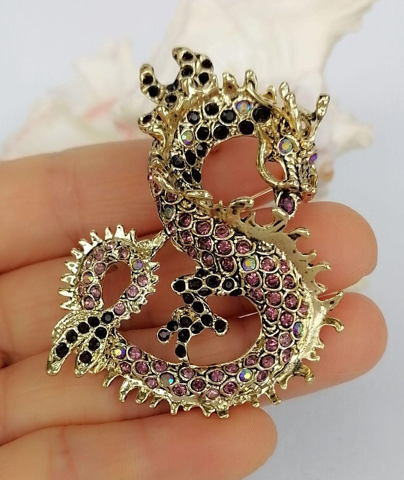 Puede incluir: Broche de drag&oacute;n dorado con pedrer&iacute;a rosa y negra. El drag&oacute;n est&aacute; intrincadamente detallado con un cuerpo curvo y detalles puntiagudos. La broche es un accesorio decorativo.