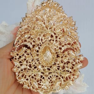May include: Ornate gold-colored brooch with intricate filigree detailing. The brooch has a central teardrop-shaped embellishment and a pin closure on the back. The design features floral and scrollwork patterns, creating a vintage or antique aesthetic.
