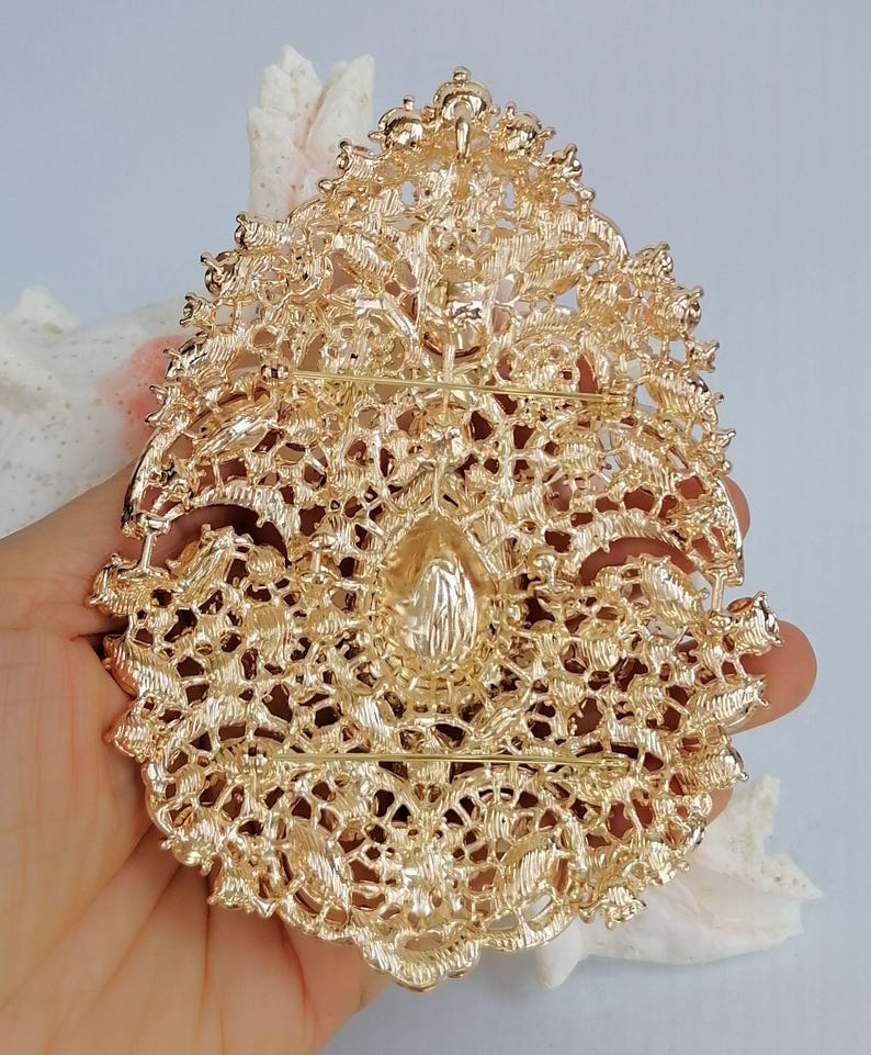 May include: Ornate gold-colored brooch with intricate filigree detailing. The brooch has a central teardrop-shaped embellishment and a pin closure on the back. The design features floral and scrollwork patterns, creating a vintage or antique aesthetic.