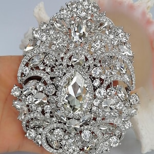 May include: An ornate, silver-toned brooch with a dazzling array of clear, sparkling crystals. The brooch features a large, pear-shaped central stone surrounded by various sizes and shapes of crystals, creating a luxurious and eye-catching design.