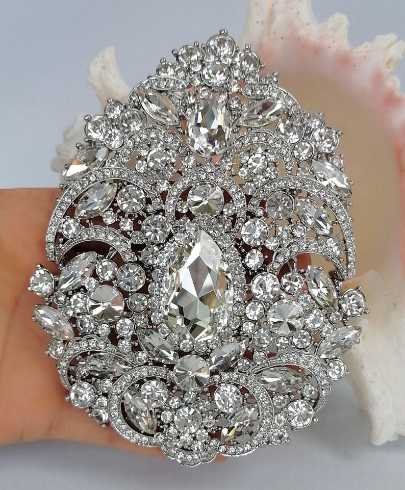 May include: An ornate, silver-toned brooch with a dazzling array of clear, sparkling crystals. The brooch features a large, pear-shaped central stone surrounded by various sizes and shapes of crystals, creating a luxurious and eye-catching design.