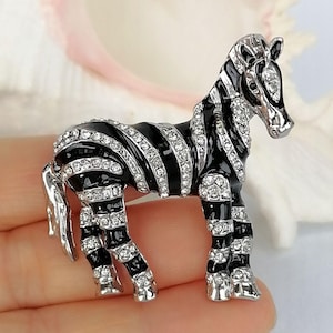 Könnte beinhalten: Zebra-Brosche mit schwarzen Emaille-Streifen und funkelnden, klaren Kristallakzenten. Das Zebra hat silberfarbene Metallbeine und einen Kopf. Dieses Schmuckstück mit Tiermotiv ist ein stilvolles Accessoire.