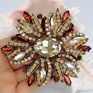Op de afbeelding: Een bloemvormige broche met een grote centrale ovale champagnekleurige kristal omringd door stralende bloemblaadjes bezet met kleine amberkleurige, bruine en heldere iriserende strass-steentjes in een donker metalen zetting.