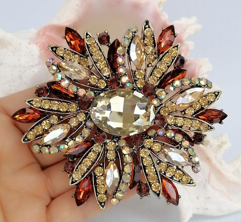Op de afbeelding: Een bloemvormige broche met een grote centrale ovale champagnekleurige kristal omringd door stralende bloemblaadjes bezet met kleine amberkleurige, bruine en heldere iriserende strass-steentjes in een donker metalen zetting.