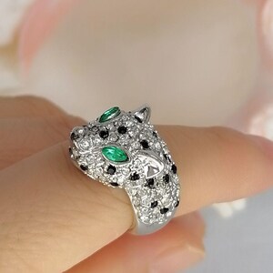 May include: A silver-toned ring shaped like a leopard head, fully encrusted with clear white crystals and small black stones. The leopard features two marquise-cut green stones for eyes, creating a striking contrast against the metallic surface.