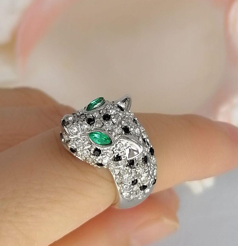 May include: A silver-toned ring shaped like a leopard head, fully encrusted with clear white crystals and small black stones. The leopard features two marquise-cut green stones for eyes, creating a striking contrast against the metallic surface.