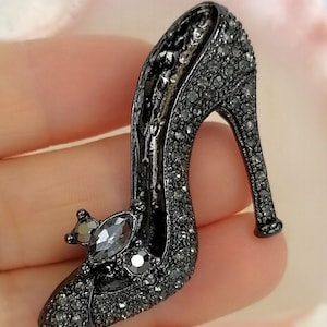May include: A black high-heeled shoe brooch, encrusted with small, dark gray rhinestones. The shoe features a pointed toe and a slender heel, with a decorative embellishment on the vamp. The brooch is a stylish accessory.