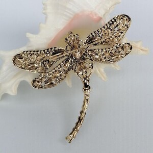 May include: A gold dragonfly brooch with detailed openwork design. The brooch is displayed on a white seashell with a pink interior. The brooch has a pin fastening on the back.