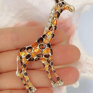 May include: A gold-toned giraffe brooch featuring an enamel pattern of orange and dark brown spots. The giraffe's neck is accented with a row of clear, sparkling rhinestones, and additional small rhinestones are set along the base of each foot.