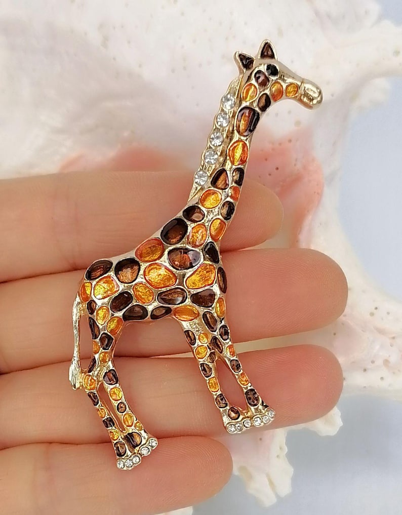 May include: A gold-toned giraffe brooch featuring an enamel pattern of orange and dark brown spots. The giraffe's neck is accented with a row of clear, sparkling rhinestones, and additional small rhinestones are set along the base of each foot.